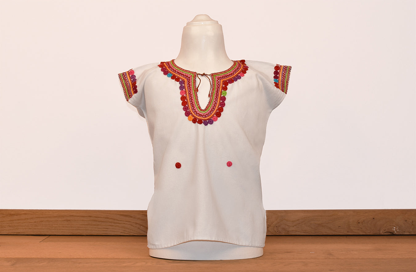 Blusa blanca para niña con bolitas y bordados de color en cuello y mangas.